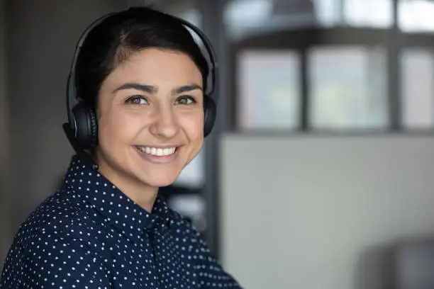 Head shot close up smiling attractive indian ethnicity millennial businesswoman wearing headphones, looking at camera. Happy sincere pleasant young mixed race female professional working remotely.