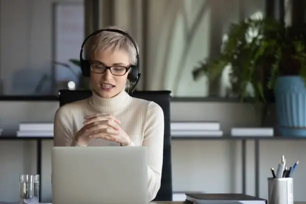 Focused pleasant 30s businesswoman in eyeglasses wearing wireless headphones with microphone, looking at laptop screen, holding video call negotiations meeting online with partners alone in office.