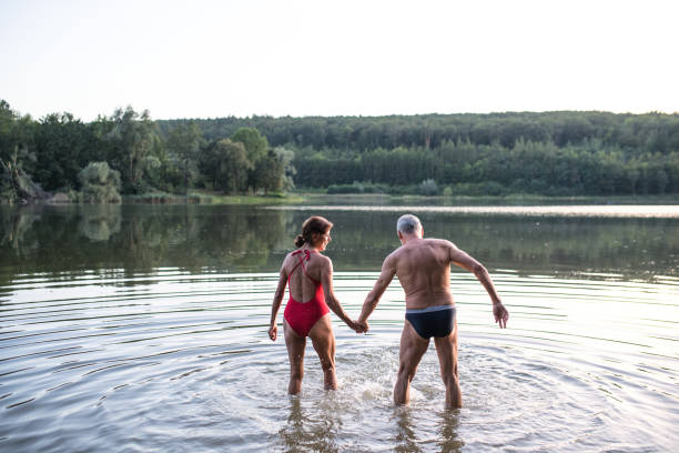 水泳前に屋外の湖に立っている水着を着たシニアカップルの背面図。 - swimming exercising women back ストックフォトと画像