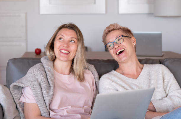 Two charming blonde women are laughing on the sofa in their cozy living room stock photo