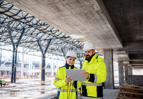 Front view of man and woman engineers standing outdoors on construction site, using tablet. Copy space.
