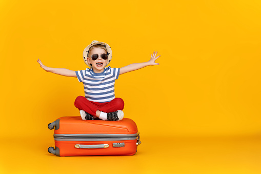 Happy little boy with outstretched arms and open mouth sitting crossed legged on luggage and pretending to fly against yellow background