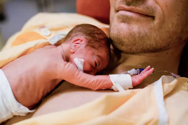 Premature baby born in the 28th week of pregnancy lies on Papa's chest and raises one hand