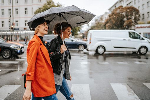 Two young female friends crossing the street on zebra crossing on rainy day in the city