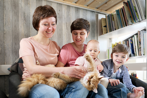A young lesbian same-sex family with two children, a son and a daughter spend time at home. They sit on the couch and read a book. The pet cat is lying next to it. Homosexual couple marriage.