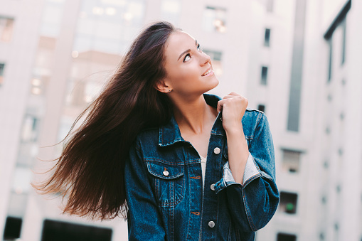 Portrait of cheerful fashionable young carefree woman posing with hair in motion outdoors. Happy beautiful trendy hipster girl looking up and smiling while walking city streets. Modern city fashion
