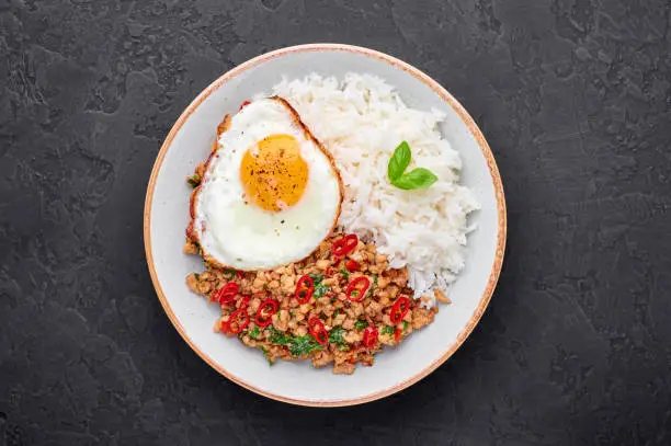Photo of Pad Krapow Gai - Thai Basil Chicken with Rice and fried Egg black slate background. Pad Krapow is Thai cuisine dish with minced chicken or pork meat, basil, soy and oyster sauces. Thai Food Copy space