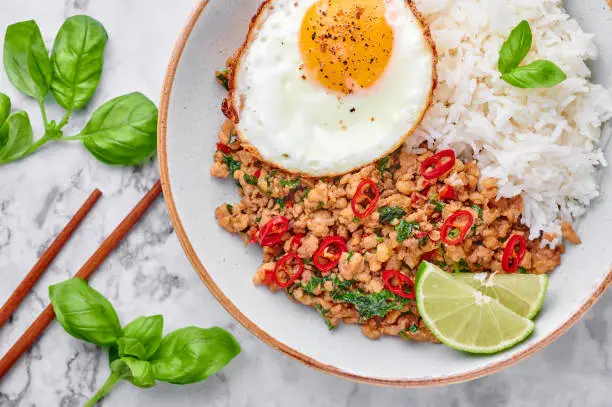 Photo of Pad Krapow Gai - Thai Basil Chicken with Rice and fried Egg on white marble background. Pad Krapow is Thai cuisine dish with minced chicken or pork meat, basil, soy and oyster sauces. Thai Food.