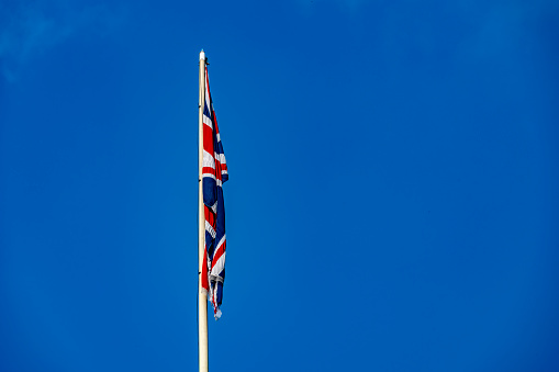 The flag of the United Kingdom of Great Britain and Northern Ireland, known as Union Flag or Union Jack, hanging down loosely at full-mast on a white pole against blue sky.