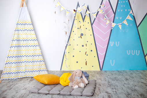 Stylish white modern kids room. Kids wigwam in childrens room. Scandinavian style of interior. Spacious bedroom for a child with a beautiful multi-colored mountain painting and pennants flags. teepee