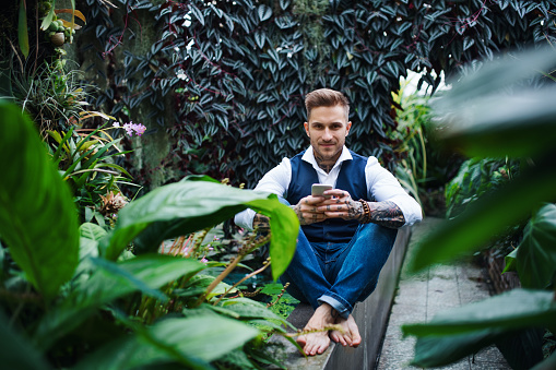 Young man sitting barefoot in botanical garden, using smartphone. Green bussiness concept.