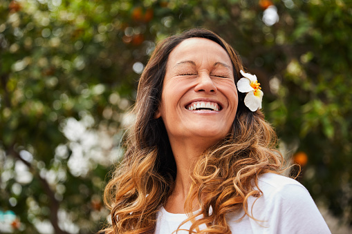 Laughing mature woman standing outside in her yard on a sunny afternoon with a flower in her long brown hair