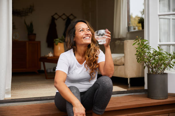 Smiling mature woman sitting on her patio drinking water Mature woman laughing while sitting alone on her patio steps outside drinking a glass of water drinks on the deck stock pictures, royalty-free photos & images