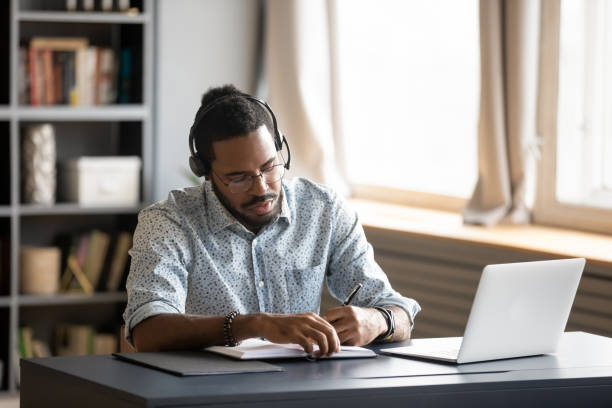 Concentrated biracial guy listening to favorite music while planning workday. Concentrated millennial african american guy wearing earphones, listening to favorite music while planning workday. Focused young biracial businessman watching educational lecture. writing down notes. business teaching student writing stock pictures, royalty-free photos & images