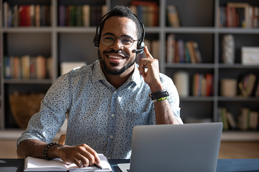 Happy young friendly african american man in eyeglasses wearing headset with mic, looking at camera. Smiling professional financial advisor consulting clients online, solving problems issues.