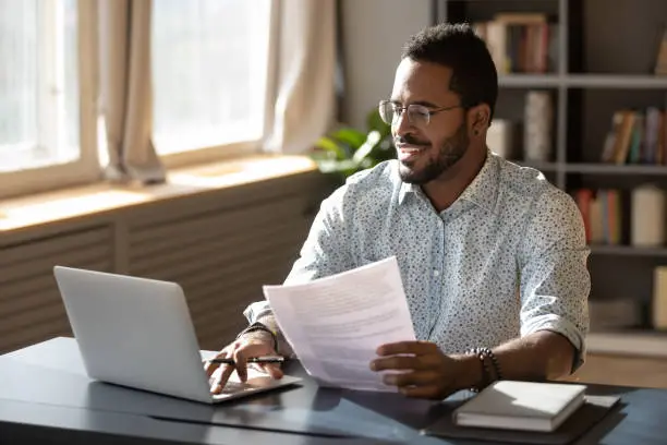 Happy millennial african american businessman in eyewear holding documents, doing paperwork, preparing report or analyzing market research results, working on computer in modern workplace office.