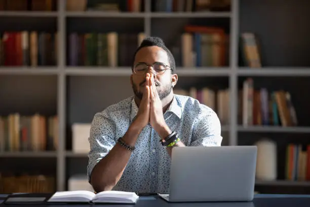 Front view frustrated millennial african american guy sitting at able with computer, praying god with folded hands, asking good luck before business start. Hopeful young biracial man making decision.