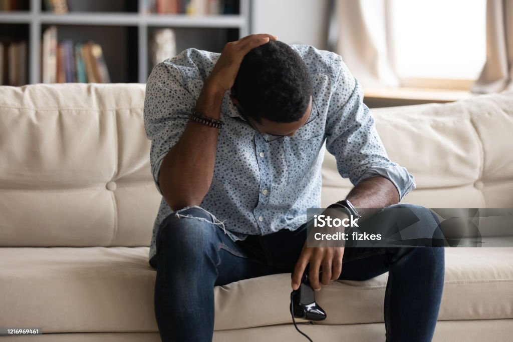 Unhappy young biracial man feeling upset after loosing xbox game. Unhappy young african american man holding joystick, feeling upset after losing xbox video game. Addicted to technology biracial guy player with gamepad controller displeased with online tournament. Video Game Stock Photo