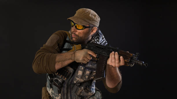 istock Soldier mercenary with a gun looking by side. Photo on a dark background. 1216970200
