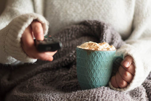 Woman covered with blanket holding tv remote and mug of hot drink with whipped cream