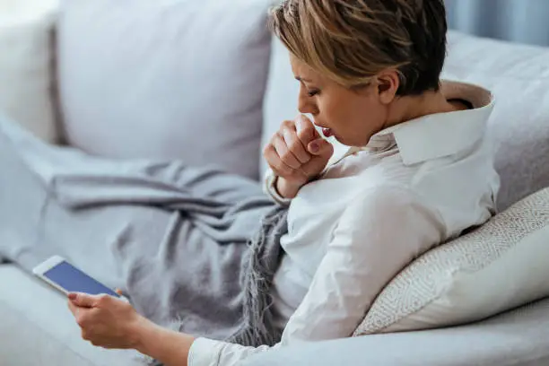 Woman having cold and flue virus and coughing while using cell phone on the sofa.