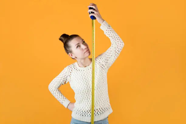 girl looks suspiciously at a measuring tape stretched next to her, isolated on orange-yellow