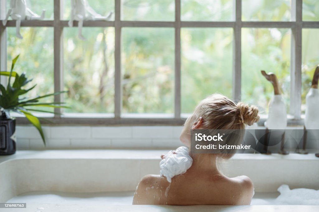 Young adult woman taking bath holding sponge in hand, washed shoulder Skin care concept. Back view of young adult woman taking bath, washed shoulder, holding sponge in hand and lying in foam water, spending morning in bathroom Bathtub Stock Photo