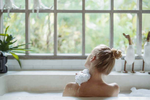 Young adult woman taking bath holding sponge in hand, washed shoulder Skin care concept. Back view of young adult woman taking bath, washed shoulder, holding sponge in hand and lying in foam water, spending morning in bathroom spa lady stock pictures, royalty-free photos & images