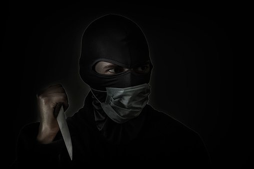 Bandit in black mask with a slit for the eyes and a medical mask waving a knife. Low hatch. Close-up. Copy space