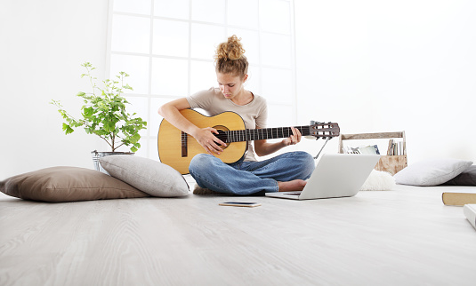 young woman sitting in living room playing guitar at the computer, stay at home concept