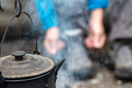 Close-up of a sooty teapot with a lid hanging and basking over a campfire, on a blurry background, a woman lacing hiking boots.