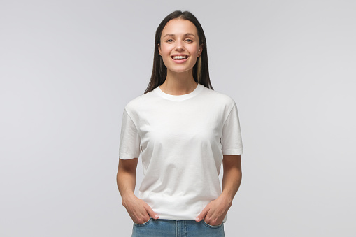 Laughing student girl wearing white T-shirt and blue jeans, standing with hands in pockets, looking straight at camera, isolated on gray background