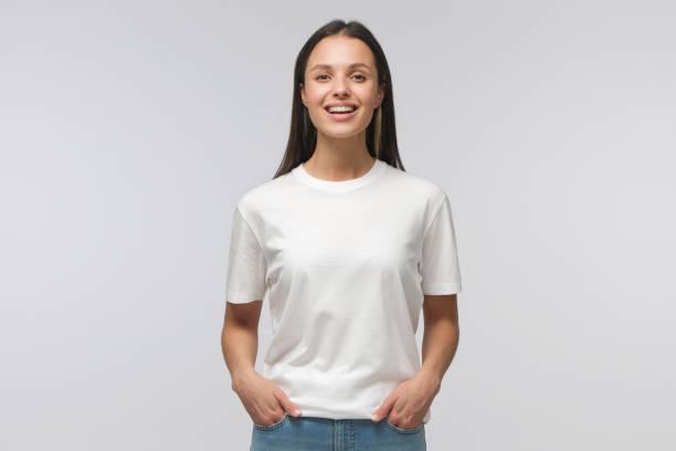 Laughing student girl wearing white T-shirt and blue jeans, standing with hands in pockets, looking straight at camera, isolated on gray background Laughing student girl wearing white T-shirt and blue jeans, standing with hands in pockets, looking straight at camera, isolated on gray background caucasian appearance stock pictures, royalty-free photos & images