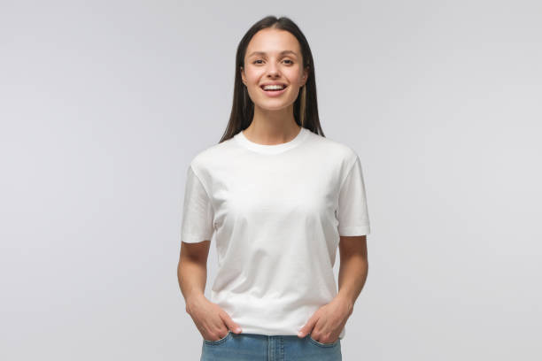 смеющаяся девушка-студентка в белой футболке и синих джинсах, стоящая с руками в карманах, глядя прямо в камеру, изолированная на сером фоне - европейского происхождения стоковые фото и изображения