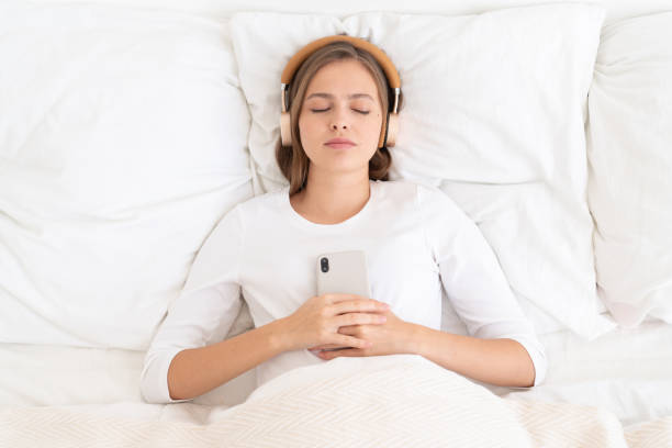 jeune fille se trouvant sur le lit blanc avec les yeux fermés le matin, retenant le smartphone, écoutant la musique de relax par des écouteurs sans fil, se relaxant - female likeness audio photos et images de collection