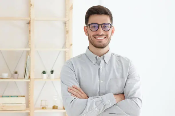 Photo of Portrait of young business man wearing gray shirt and glasses, stand with crossed arms in office, looking confident, professional, smiling positively at camera