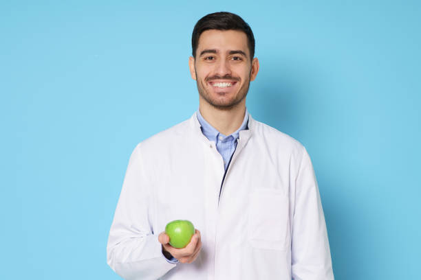 白いコートと丸い眼鏡をかけ、緑色のリンゴを手に持ち、青い背景に隔離された笑顔の栄養士または歯科医の医師 - food white caucasian color image ストックフォトと画像