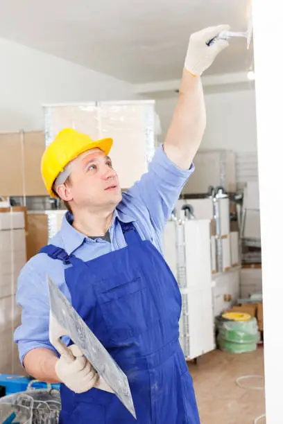 Construction worker wearing coverall and hard hat renovating walls in room with plastering tools