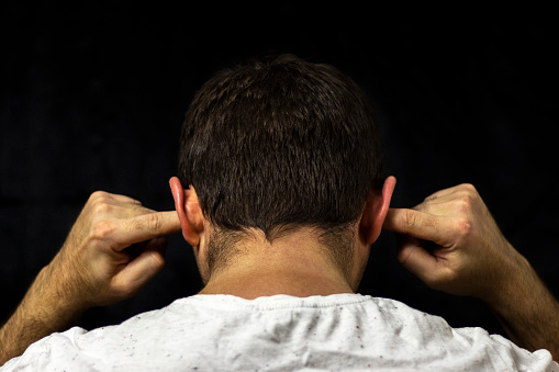 A man in a white T-shirt covers his ears with his fingers. Close up on black background. Back view.