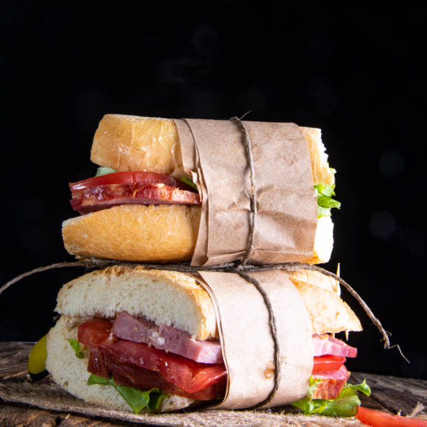 Two sandwiches with meat, tomatoes and salad in a paper wrapper, on a linen napkin. Black background. Two sandwiches with meat, tomatoes and salad in a paper wrapper, on a linen napkin. Side view. Black background. тост stock pictures, royalty-free photos & images