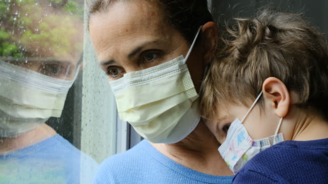 Mature woman posing with her son, both with protective masks, very sad looking through window worried about Covid-19 lockdown
