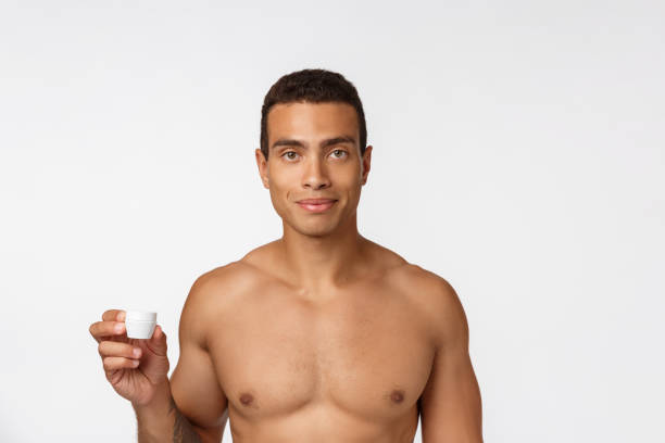 foto di un uomo afro-americano a torso libero che sorride e applica crema per il viso isolata su sfondo bianco - hairy men shaving chest foto e immagini stock