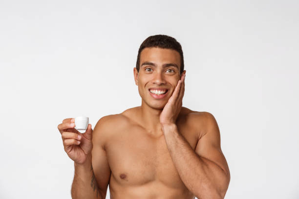 foto di un uomo afro-americano a torso libero che sorride e applica crema per il viso isolata su sfondo bianco - hairy men shaving chest foto e immagini stock