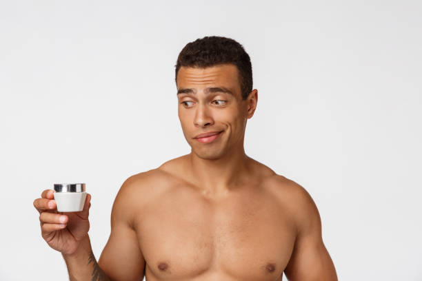 foto di un uomo afro-americano a torso libero che sorride e applica crema per il viso isolata su sfondo bianco - hairy men shaving chest foto e immagini stock