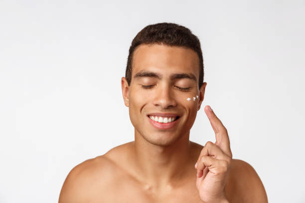 foto di un uomo afro-americano a torso libero che sorride e applica crema per il viso isolata su sfondo bianco - hairy men shaving chest foto e immagini stock