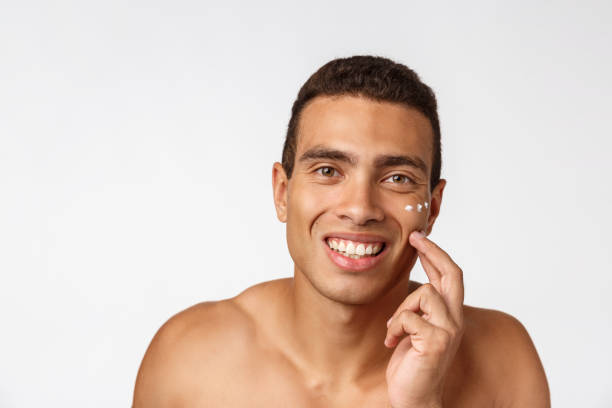 foto di un uomo afro-americano a torso libero che sorride e applica crema per il viso isolata su sfondo bianco - hairy men shaving chest foto e immagini stock