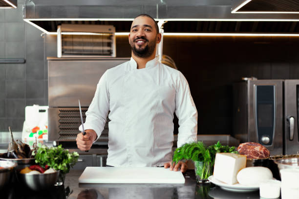 istock Handsome young African chef standing in professional kitchen in restaurant preparing a meal of meat and cheese vegetables. 1217012850