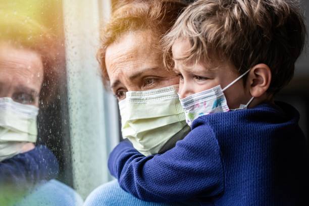 Mature woman posing with her son, both with protective masks, very sad looking through window worried about Covid-19 lockdown
