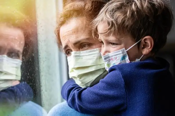 Caucasian Mature woman posing with her son, both with protective masks, very sad looking through window worried about Covid-19 lockdown