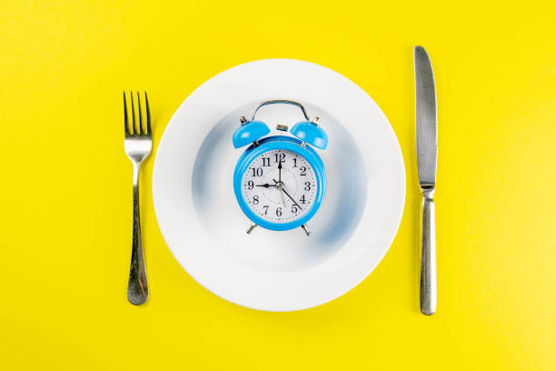 istock Alarm clock with fork and knife on the white plate on yellow table. Time to eat, Breakfast, Lunch Time and Dinner concept 1217020531